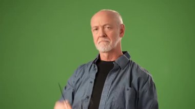 Sakallı olgun bir adam, yeşil bir arka fonun önünde dikilirken gözlüğünü düşünceli bir şekilde tutuyor. Portresi, derin bir özgüven ifadesi sergiliyor, merak uyandırıyor..