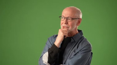 Gözlük takan olgun bir adam düşünceli bir şekilde yeşil ekran arkaplanına karşı durarak ifadesindeki derin düşünce ve duyguyu çağrıştırıyor..