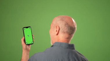 Olgun bir adam elinde akıllı telefonuyla yeşil ekran karşısında ürün duyurusu yaparken gülümsüyor. Odak noktası onun olumlu duyguları..