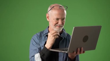 Gri saçlı ve gözlüklü olgun bir adam, ürün anonsları için katı yeşil ekran arka planına karşı dizüstü bilgisayarını düşünceli bir ifadeyle inceliyor..