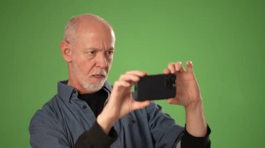 Gri saçlı ve sakallı olgun bir adam, yeşil ekrana karşı akıllı telefonuyla fotoğraf çekmeye odaklanır. Duygularını ve nişanını ifade eder..