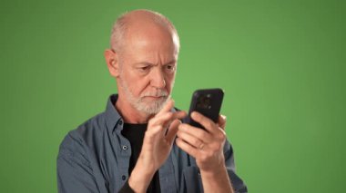 Gri saçlı olgun kel bir adam akıllı telefonuyla iletişim kuruyor ve hayal kırıklığı ve öfke gösteriyor. Katı yeşil ekran arkaplanında izole edilmiş.