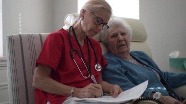 Kırmızı önlüklü bir sağlık görevlisi, rahat bir sandalyede oturan yaşlı bir kadına yardımcı olur. Sağlığını ve rahatlığını sıcak bir ortamda sağlarken not alır..