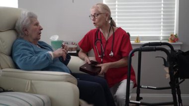 Kırmızı üniformalı bir sağlık çalışanı, rahat bir sandalyede oturan yaşlı bir kadınla yakınlaşıyor ve ev ziyareti sırasında şefkatli bir atmosfer yaratıyor..