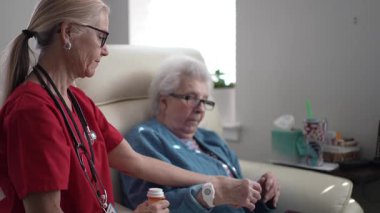 Kırmızı önlüklü bir sağlık görevlisi rahat bir sandalyede oturan yaşlı bir kadına yardım ediyor. Parlak bir odada etkileşim sıcak ve destekleyicidir..