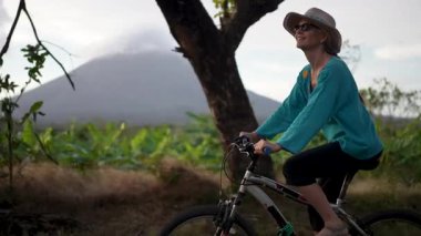 Gülümseyen olgun bir kadın Ometepe Adası 'nda verimli bir çevrede bisiklet sürmekten zevk alırken arka planda muhteşem Concepcion volkanı Orta Amerika' da doğal güzellikleri gözler önüne seriyor..