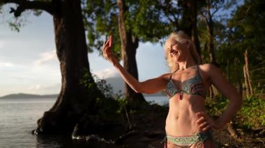 Gülümseyen olgun bir kadın telefonu ile Nikaragua 'daki sakin gölün yanında neşeli bir selfie çekiyor. Etrafı doğa ve çiçek açan ağaçlarla çevrili. Orta Amerika' da yaz tatilinin tadını çıkarıyor..
