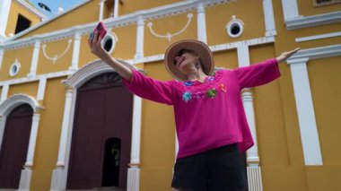 Seyahatinin keyfini çıkaran olgun bir kadın, Moyogalpa, Ometepe Adası 'ndaki canlı bir kilisenin önünde neşeli bir selfie çekiyor ve turizmin ortasında neşeli ruhunu gösteriyor..