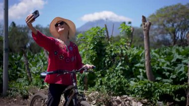 Gülümseyen son sınıf öğrencisi bir kadın Ometepe Adası 'nda bisiklet sürmekten hoşlanıyor. Güneşli bir günde, Volkan Concepcion' un arka planında telefonuyla fotoğraf çekiyor..