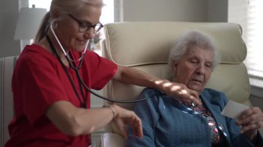 Kırmızı önlüklü bir hemşire rahat bir şekilde oturan yaşlı bir kadının kalbini dinliyor. Aydınlık ve rahat bir ortam, özen ve merhameti ön plana çıkarıyor..