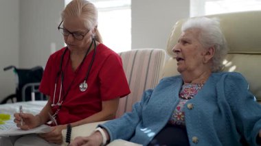 Kırmızı önlüklü bir sağlık görevlisi, rahat bir yaşam alanında yaşlı bir kadınla ilişki kurarken not alıyor..