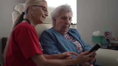 Bir sağlık çalışanı, yaşlı bir kadına uzaktan kumandayla ev ortamında rehberlik eder. Etkileşimde yaşlılar için teknoloji desteği rahatça otururken vurgulanıyor.