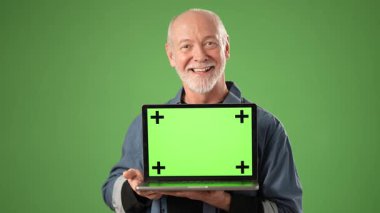 Kıdemli adam yeşil arka planda duruyor, elinde yeşil ekranlı bir laptop tutuyor, yaratıcı projeler ve video düzenleme alanı için hazır..