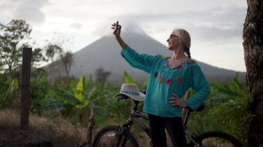 Renkli bluzlu mutlu bir son sınıf öğrencisi Ometepe Adası 'nın arka planında yemyeşil çimlerle çevrili Concepcion volkanı ile neşeli bir selfie çeker..