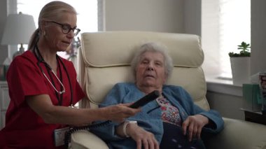 Bakıcı, yaşlı bir kadının rahat bir koltukta otururken uzaktan kumanda kullanmasına yardım eder. Doğal ışık, sıcak odayı doldurur ve sıcak bir atmosfer yaratır..