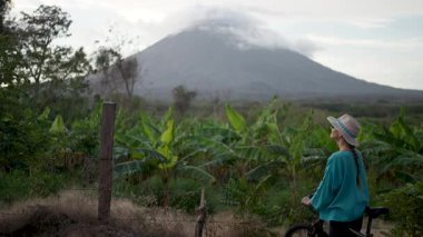 Güzel, gülümseyen, olgun bir kadın motosikletinin yanında duruyor, Ometepe Adası 'nın bereketli manzarasına ve Orta Amerika' da bulutlu bir gökyüzünün altındaki görkemli Concepcion volkanına hayran..