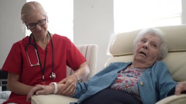 Bir bakıcı yaşlı bir kadının koluna nazikçe masaj yapar, öğleden sonra saatlerinde aydınlık bir klinik ortamda rahatlık ve konfor sunar..