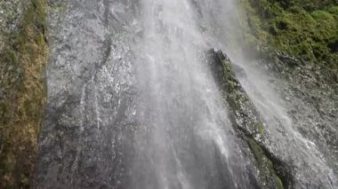 Kameralar, olgun yaşlı bir kadının Nikaragua, Ometepe 'deki çağlayan bir şelalenin altında, yemyeşil ormanlarla çevrili olduğunu gösteriyor. Güneş ışığı spreyde bir gökkuşağı yaratır..