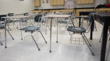 Masalar ve sandalyeler boş bir sınıfta, öğrencilerin gelip öğrenme faaliyetlerine girişlerini düzgünce düzenliyorlar. Pencerelerden güneş ışığı akıyor..