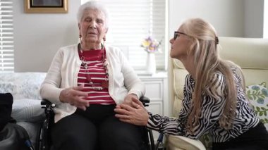 90 'lı yaşlarda yaşlı bir kadın tekerlekli sandalyede otururken kızı nazikçe elini tutuyor sıcak ışıklarla dolu sıcak oturma odalarında şefkatli bir anı paylaşıyor..