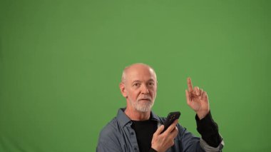 Olgun bir adam, yukarı doğru işaret ederken ve akıllı bir telefon tutarken heyecan gösterir. Sağlam yeşil bir arka planı vardır. Ürün duyuruları için ideal bir kopya alanı yaratır..