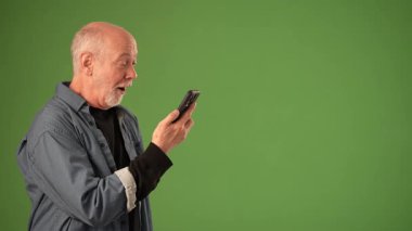 Olgun bir adam, yeşil ekranın önünde dikilirken akıllı telefonu zevkle tutar ve işaret eder. İfadesi bilgi paylaşımında heyecan ve coşku gösteriyor..