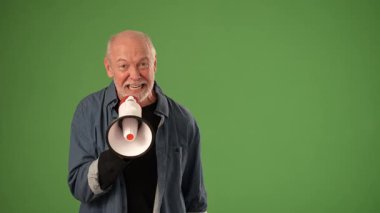 Gri saçlı olgun bir adam yeşil perde arkasında tutkulu bir şekilde megafona konuşuyor. Ürün duyurusu ya da kutlama için güçlü duygular ifade ediyor..