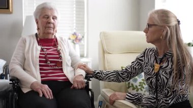 Yaşlı bir kadın tekerlekli sandalyede otururken kızı nazikçe koluna dokunuyor. İyi aydınlatılmış bir oturma odasında sessiz bir anı paylaşıyorlar..