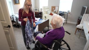 Parlak, rahat bir oturma odasında, bir kız, yaşlı annesine tekerlekli sandalyede otururken yeni bir kıyafet gösterir, neşe ve bağlantı anlarını paylaşır..