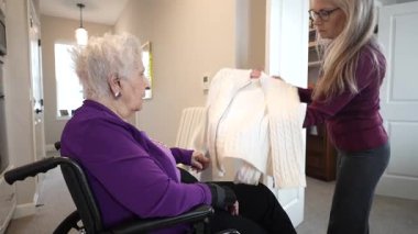 Tekerlekli sandalyedeki yaşlı bir kadın sıcak bir oturma odasında rahat bir kazak seçerken kızından yardım alıyor..