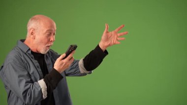 Olgun bir adam akıllı telefonuyla neşeli bir şekilde etkileşime girer ve sağlam yeşil bir zemin üzerinde coşkuyla işaret eder. Onun ifadesi şaşkınlık ve mutluluk taşıyor..