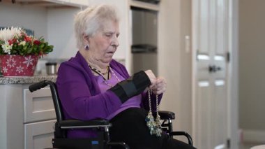 Mor hırka giymiş yaşlı bir kadın evde tekerlekli sandalyede oturuyor. Mücevher tokasını açmaya çabalıyor, rahat ortamında yaratıcı bir anın tadını çıkarıyor..