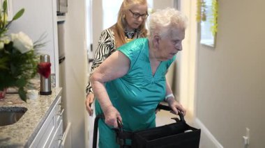 Bir aile üyesi, aydınlık ve rahat bir yerde günlük aktiviteler sırasında yürüteç kullanarak, bakım ve destek göstererek, yaşlı bir kadının evini dolaşmasına yardımcı olur..