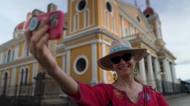 Olgun bir kadın, Granada, Nikaragua 'nın canlı mimarisinin önünde selfie çekiyor. Seyahat deneyiminin ve yerel kültürün tadını çıkarıyor..