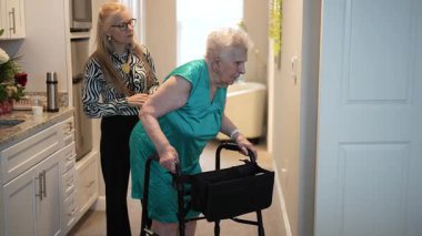 90 'lı yaşlarda yaşlı bir kadın, parlak ve davetkar bir şekilde kızlarıyla yürüteç kullanıyor. Sıcak bir ortamda bir anlık bakımı paylaşıyorlar..