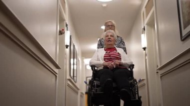 Columbia, MD, ABD - 12252024: Tekerlekli sandalyedeki yaşlı bir kadına, yakın bağlarını ve desteklerini sergileyerek, parlak bir koridorda ilerleyen kızı yardımcı oluyor.