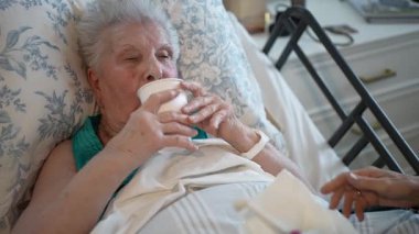 90 'lı yaşlarda yaşlı bir kadın, yumuşak bir fırça kullanarak biri ona diş hijyeninde yardım ederken yatakta dinleniyor. Rahat ve bakımlı görünüyor..