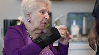 Bileklik takan yaşlı bir kadın, ilaçlarını zarif dekorlarla çevrili, kapalı bir ortamda içerken dikkatlice su içer..