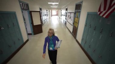 Gözlüklü orta yaşlı bir kadın okul koridorunda dolap ve Amerikan bayrağıyla birlikte yürürken kitap ve öğretim malzemeleri taşıyor..