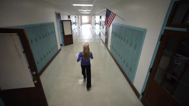 Gündelik kıyafetler giyen olgun bir kadın okul koridorunda dolaplarla dolu bir şekilde yürüyor. Yukarıda bir Amerikan bayrağı asılı, eğitici atmosferi geliştiriyor..