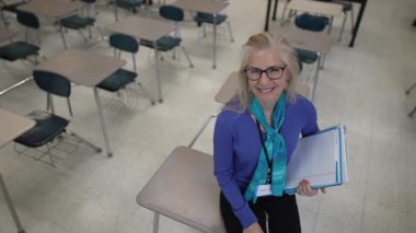 Orta yaşlı bir kadın boş bir sınıfın masasına oturmuş elinde bir not panosuyla içten bir şekilde gülümsüyor. Öğretmek için özgüven ve coşku yayıyor..