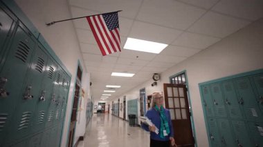 Orta yaşlı, gözlüklü bir kadın okul koridorunda beklerken elinde kağıtlar tutuyor. Metal dolaplar duvarlarda asılı ve yukarıda bir Amerikan bayrağı asılı..