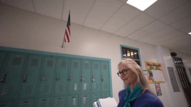Orta yaşlı bir kadın öğretmen elinde kitapla okul koridorunda yürüyor. Parlak renkli dolaplar asılı bir Amerikan bayrağının altında duvarları sarıyor..