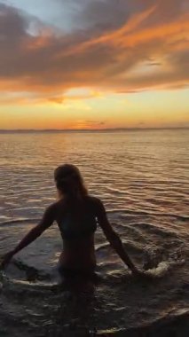 Orta yaşlı, gülümseyen mutlu bir kadının dikey video silueti gün batımında Orta Amerika gölünün sakin sularında duruyor. Tatilinin güzelliğini kucaklayarak neşe saçıyor..