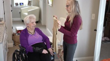 Bir bakıcı, tekerlekli sandalyedeki yaşlı bir kadına yardım eder. Etrafı kişisel eşyaları ve güneş ışığını içeren rahat bir ev ortamıyla çevriliyken kıyafetleri tartışır..