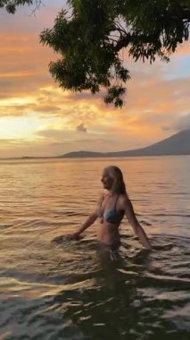 Mutlu, gülümseyen, olgun bir kadının günbatımında dinlendirici sularda yürüyen, tatilinde okyanusun ya da gölün sükunetini kucaklayan dikey video portresi..