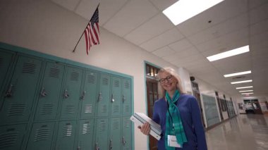 Orta yaşlı bir kadın öğretmen elinde birden fazla dosya tutarken okulun koridorunda gülümsüyor. Dolaplar duvarlara dizilmiş ve başınızın üstünde bir Amerikan bayrağı asılı. Hoş geldin atmosferi yaratıyor..