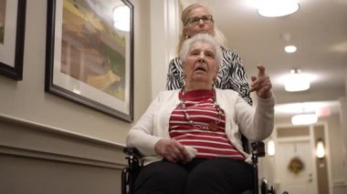 Kızı onu iterken tekerlekli sandalyedeki yaşlı bir kadın, sanatla aydınlık bir bakım evinin koridorunda neşeli bir anı paylaşıyor..