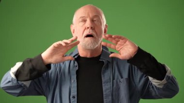 Üst düzey bir adam, katı yeşil bir altyapının önünde duruyor, açık elleri ve ifadesel yüzüyle çeşitli duygular sergiliyor, izleyiciyi doğrudan kendine çekiyor..