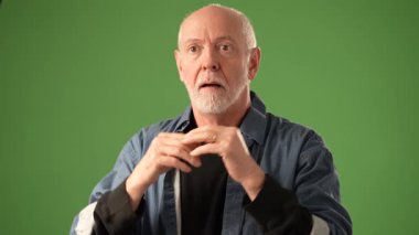 Gözlük takan olgun bir adam düşünceli bir şekilde yeşil ekran arkaplanına karşı durarak ifadesindeki derin düşünce ve duyguyu çağrıştırıyor..